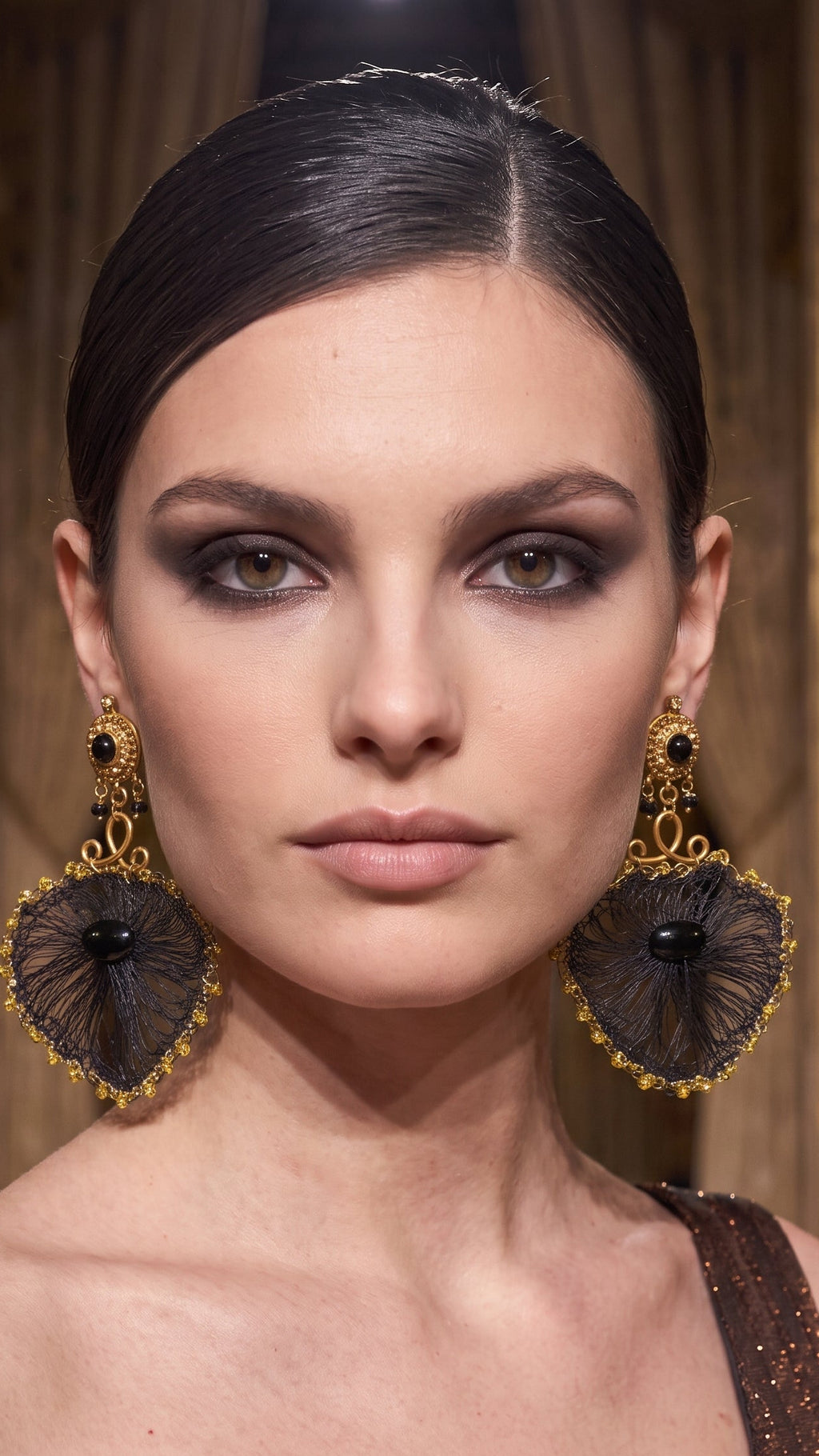 A close-up portrait of a model wearing large, ornate gold and black dangle earrings with a fan-like black mesh design and gold filigree borders.