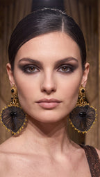 A close-up portrait of a model wearing large, ornate gold and black dangle earrings with a fan-like black mesh design and gold filigree borders.